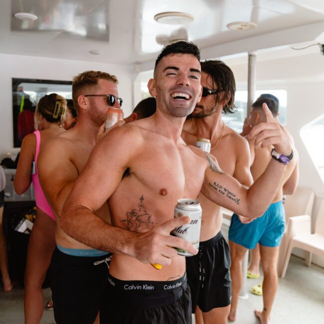 A shirtless man wearing black Calvin Klein shorts smiles and gestures while holding a beverage can on a boat. He is surrounded by other casually dressed people, one wearing a bright pink swimsuit, enjoying a lively social gathering. The setting appears casual and relaxed under the clear blue sky.