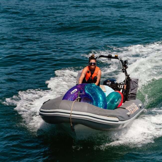 A person wearing an orange life jacket drives a small inflatable motorboat on a body of water. Several colorful inflatable rings are piled in the front. Logos reading "Sail Gustavo" and "Yacht Social Club" are visible in the corners of the image, adding to the leisure scene.