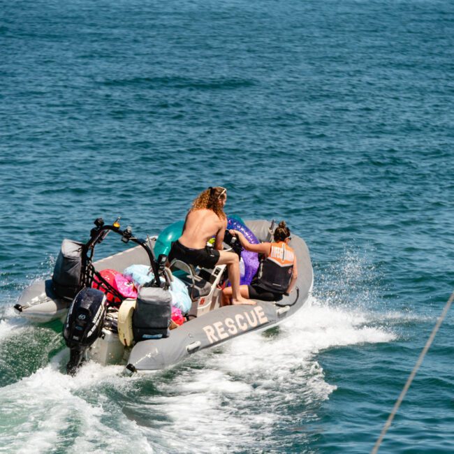 A small inflatable rescue boat with three people and equipment on board speeds across a calm blue sea. The boat, equipped with outboard motors, has one person at the front, another steering, and a third holding onto essential gear.