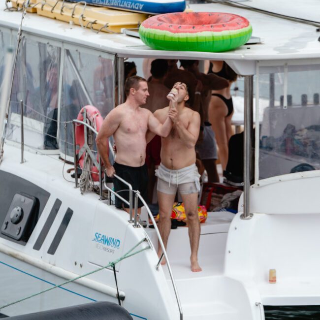 Two men stand on the deck of a boat named "Seawind." Both are shirtless, one in black shorts and the other in white shorts, sipping from a can. In the background, there's a watermelon float and other people enjoying their time on the boat.