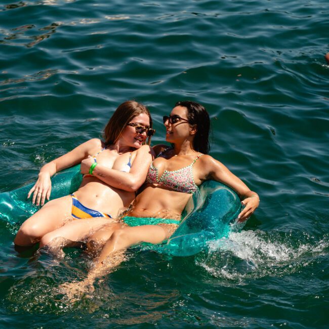 Two people relax on a blue inflatable float in a shimmering body of water, smiling and enjoying the sunny day. Others are swimming in the background, while sunlight dances on the water's surface. Logos "Sail Gustavo" and "The Yacht Social Club" can be seen.