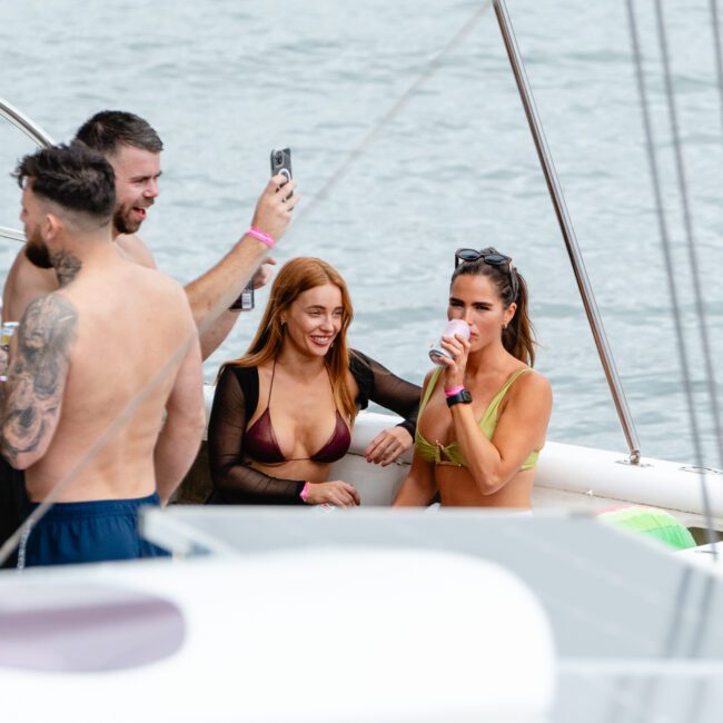 A group of people are socializing on a boat. Two women in bikinis are in the foreground, one with a tattoo sleeve and another holding a drink. A man with tattoos is capturing the moment with his smartphone. The boat sails on calm waters, creating a relaxed and fun atmosphere thriving with good vibes.