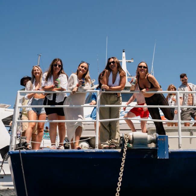 A group of people stand and lean against the railing on the upper deck of a private yacht charter in Sydney Harbour, smiling and posing for a photo. Other boats are visible in the background under a clear blue sky.