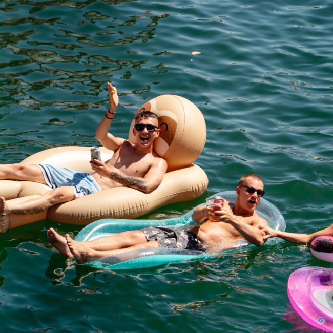 Two men are lounging on inflatable pool floats in the water, holding beverages and waving at the camera. They appear relaxed and are enjoying their time in the sun. The water is a deep green shade, with logos saying "Sail Gustavo" and "Uplift Social" visible in the bottom corners.