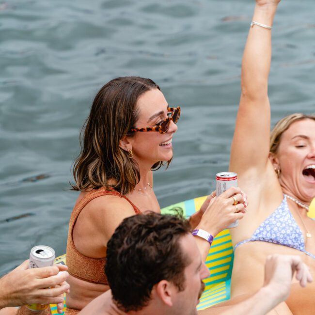Two women in swimwear and sunglasses, and a man in swim trunks, relax on inflatable rafts in the water. The woman on the left holds a beverage can and smiles, while the woman on the right raises her arm in excitement. The man also holds a drink, all enjoying their time under the sun.