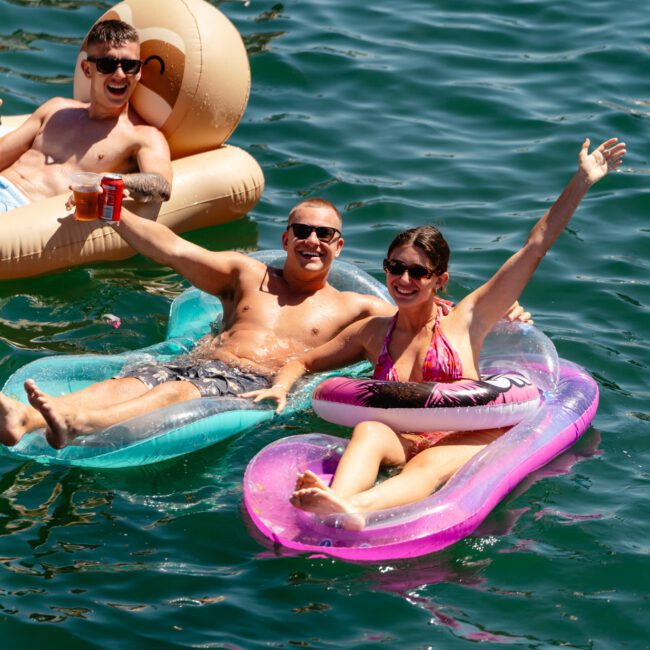 Three people smile and relax on inflatable floats in the water. One person holds a red cup while sitting on a pretzel-shaped float, while two others share a vibrant blue and pink float, raising their arms in joy. The water is a deep green. Logos read "Sail Gustavo" and "Yacht Social Club.