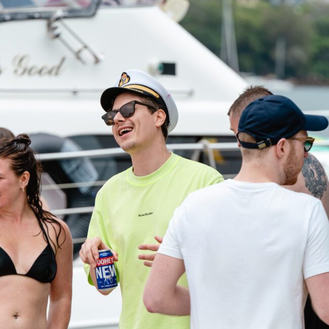 A group of people are gathered on a boat. A man wearing a neon green shirt, captain's hat, and sunglasses is laughing and holding a can of beer. Other people in swimsuits are talking and enjoying the sunny day on the water. A yacht with the name "Life's Good" is in the background.