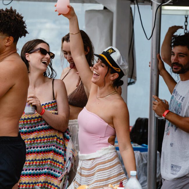 A lively group of people enjoys a party on a boat. One woman, wearing a captain's hat and a pink swimsuit, stands out as she raises her tropical drink and smiles. Others around her laugh and chat, with some also holding drinks. The atmosphere is festive and joyful.