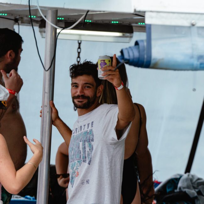 A man stands under a canopy with a beverage in hand, smiling at the camera. Dressed in a casual t-shirt, he's amidst others who are also casually dressed, likely enjoying their time on a boat or poolside. Other people are seen relaxing in the background.