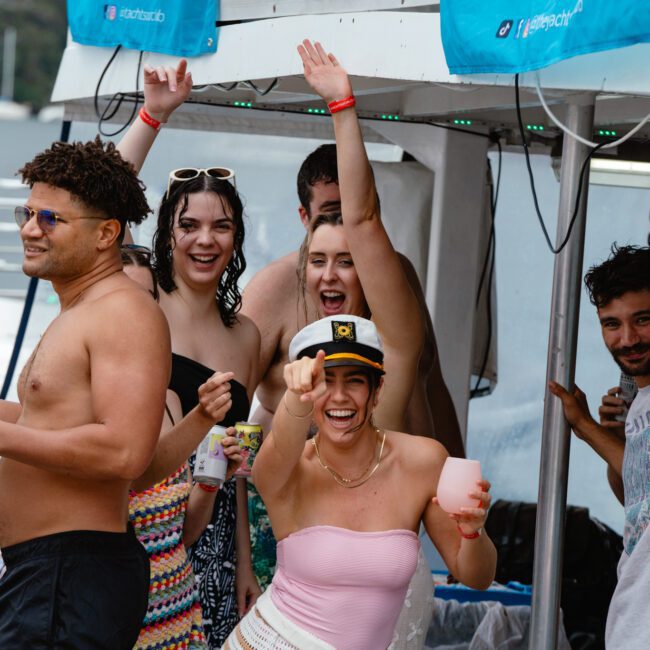 A group of friends in swimsuits are enjoying a lively boat party. One person in a captain's hat holds a drink and raises an arm in excitement. Others around them are smiling, raising their arms, and holding drinks, while the boat is adorned with festive decorations and fairy lights.