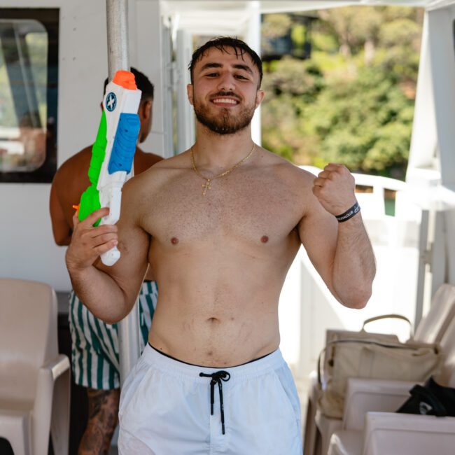 A man stands on a boat holding a colorful water gun, smiling and raising one arm in a celebratory gesture. He is shirtless and wearing white shorts. Another person is partially visible in the background, with lush trees and greenery in the distance.