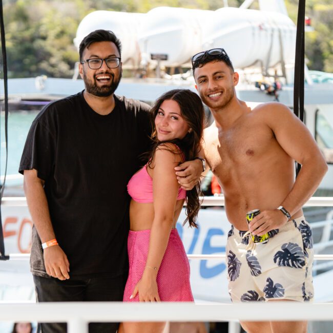 Three people are smiling and posing together on a boat. The one on the left dons a black T-shirt, the center one shines in a pink outfit, and the right person is shirtless in blue patterned swim trunks. The background beautifully showcases other boats and abundant greenery, emphasizing their joyful outing.