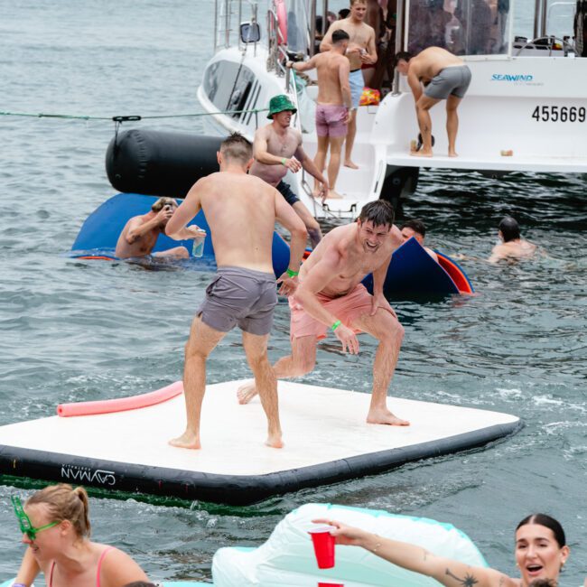 A lively scene of people enjoying themselves on a picturesque lake. Two men, each holding a pool noodle, are engaged in playful combat while standing on a floating platform. Others are swimming, lounging on inflatables, or socializing on and around sailboats in the background.