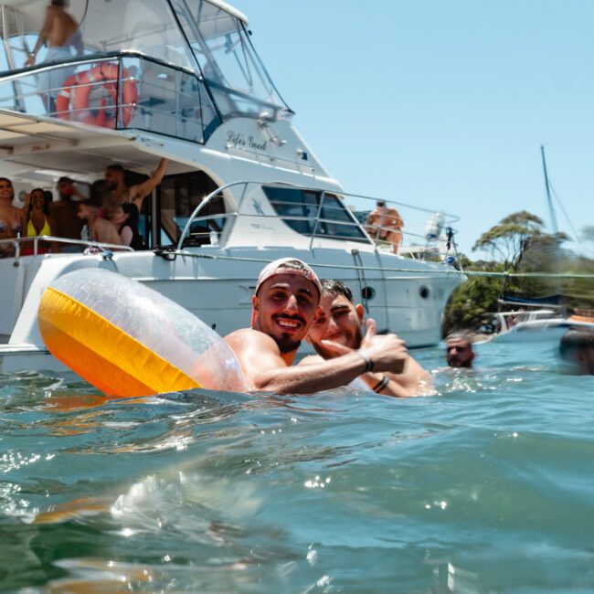 People are enjoying a sunny day in the water near a boat. In the foreground, two individuals smile and give a thumbs up, one holding an inflatable tube. Others relax and socialize on the boat in the background. The scene highlights the joy of sailing with Sail Gustavo & Get Social.