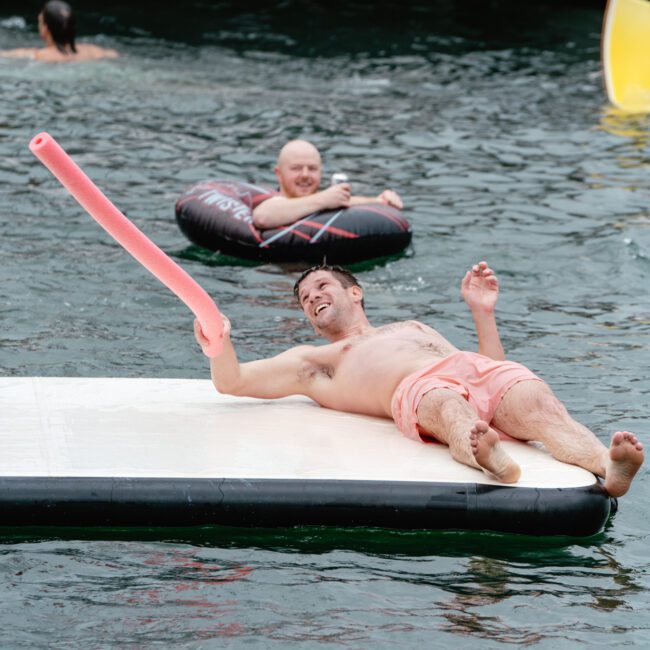 A shirtless man in pink shorts lounges on an inflatable platform in a body of water, playfully holding a pool noodle. Another person floats nearby in an inner tube, while others swim and relax. A luxurious yacht can be seen in the background.