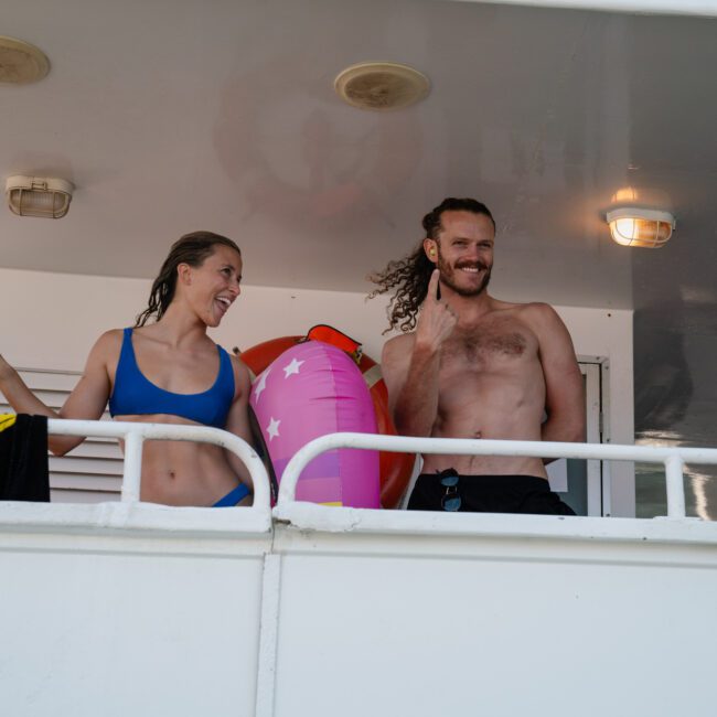 A man and a woman on a boat deck in Sydney smile at the camera, with the man waving. Both are wearing swimwear, and there is a pink inflatable ring and a yellow towel visible in the background. It looks like they're enjoying a catamaran party Sydney style!