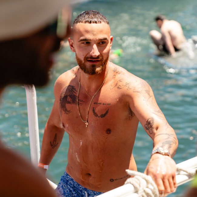 A man with tattoos on his chest and arm, wearing a blue swimsuit, stands near the edge of a boat, holding a rope and looking ahead. Other people are swimming and relaxing in the water in the background. The scene appears to be sunny, lively, and full of excitement.