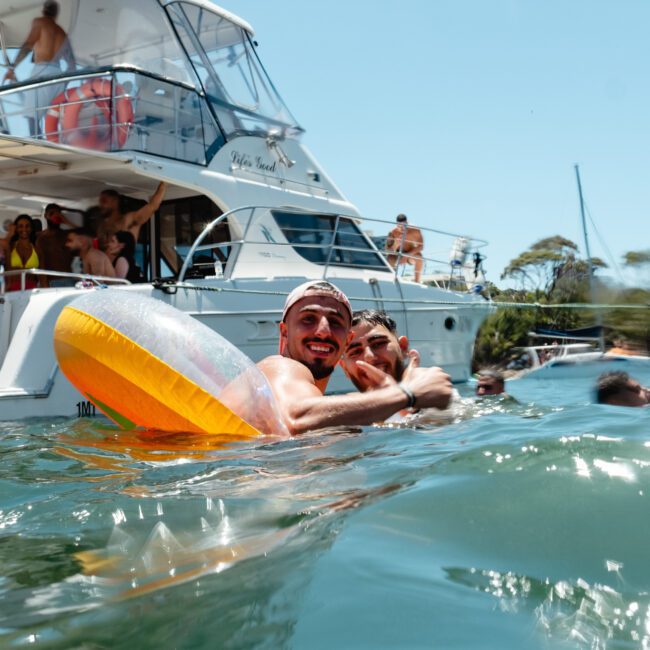 Two people smile and pose for a photo while swimming near a luxurious yacht. One is holding a colorful inflatable ring. In the background, people are relaxing on the yacht and enjoying the sunny day. The water is calm and clear, reflecting the bright sky perfectly.