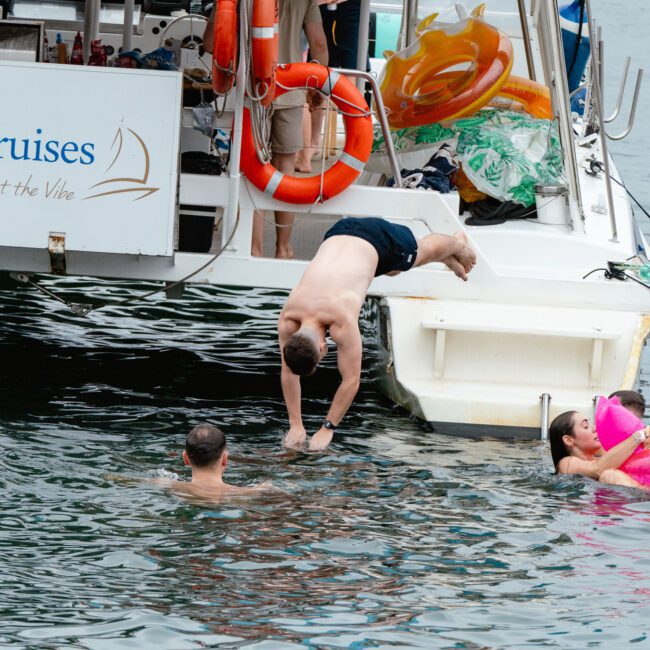 A man is diving off a boat labeled "Runner Cruises" into the water, while another man swims nearby. A woman lounges on a pink inflatable unicorn to the right. The boat's deck shows scattered life vests and inflatables, adding to the fun atmosphere.