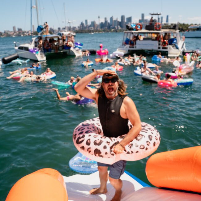 A man wearing a leopard-print swim ring stands on a float in a crowded water area with boats and people in the background, while city buildings are visible in the distance. A DJ boat hire Sydney event adds to the lively atmosphere.