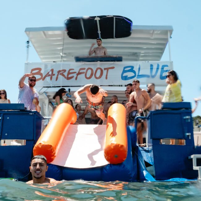 A group of people enjoys a day on a boat named "Barefoot Blue." Some are on the boat, while others are sliding off an orange slide into the refreshing water. Everyone is dressed in swimwear and appears to be having a fantastic time under the sunny weather.