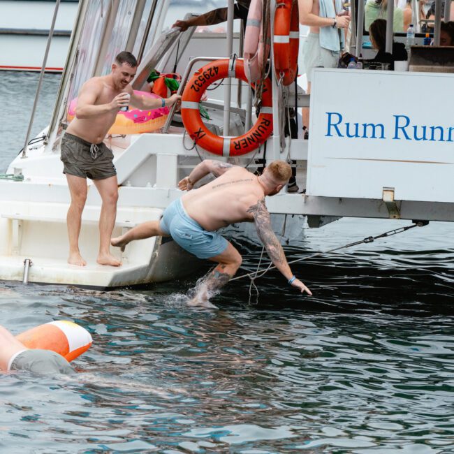 Several people are having fun on and around a Rum Runner Cruises boat. Some are in the water, and one person is mid-jump off the boat. Two men are about to jump with one holding a toy water gun, adding excitement to the scene. A life ring is attached to the side of the boat.