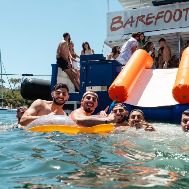 A group of young men enjoys swimming near a party boat named "Barefoot Blue." Some are in the water, one with a yellow float, while others are on the boat. The boat boasts an inflatable water slide and vibrant music. The atmosphere is lively and sunny.