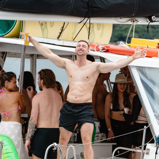 A shirtless man in black shorts and sandals stands with arms outstretched on a boat, surrounded by people in swimsuits. A bridge and trees are visible in the background. The boat is filled with summer items like inflatable toys and has a casual, festive vibe.
