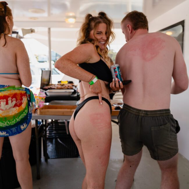 A group of people on a boat, with one woman in a black bikini pointing to her lower back, smiling at the camera. Another shirtless man has a sunburn on his back and is holding a can. It looks like they’re enjoying their time on a private yacht charter Sydney Harbour.