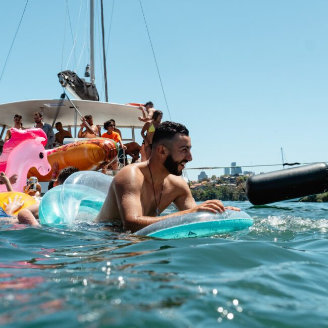 A group of people enjoying a sunny day on the water, with several individuals floating on inflatable pool toys near a boat. One man in the foreground is smiling on an inflatable lounger. The background shows more people on the boat and a picturesque city skyline reflecting off the serene surface.
