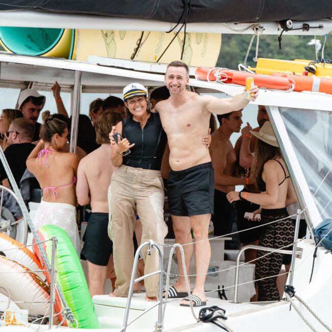 A group of people is gathered on a boat, socializing and enjoying a sunny day. Two people in the foreground, one wearing a hat and sunglasses and the other shirtless, smile and pose for the camera. The boat is vibrantly decorated with colorful pool floats.