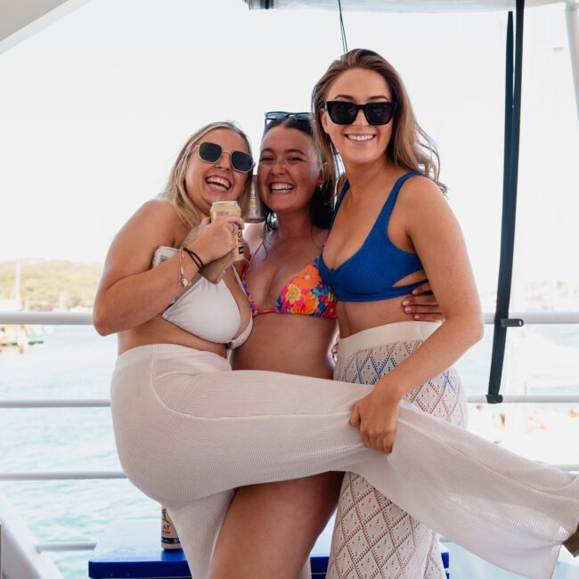 Three women, wearing swimsuits and sunglasses, stand on a boat deck smiling and posing for a photo. The background shows a scenic view of water and distant shorelines, capturing the essence of luxury yacht hire Sydney.