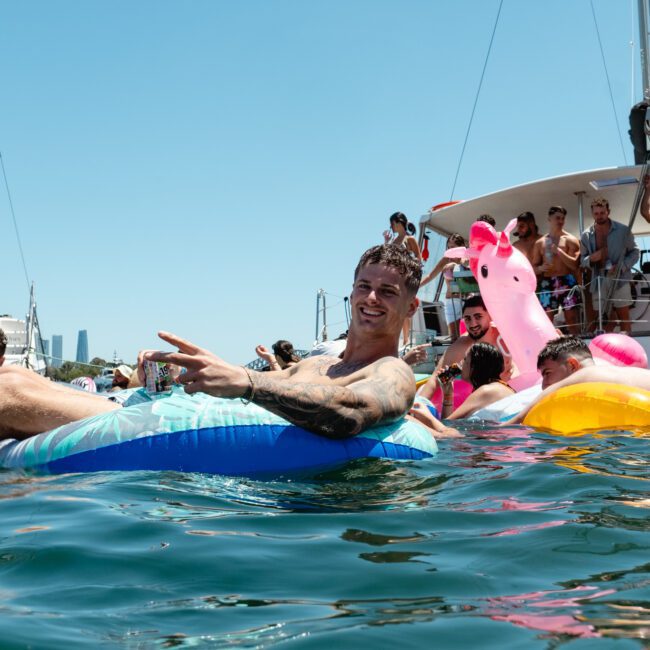 A man is lying on a float in the water, making a peace sign. Other people are floating nearby on various inflatables, including an impressive rainbow-colored unicorn. A boat with more people on deck is in the background. The atmosphere is festive under a clear blue sky.