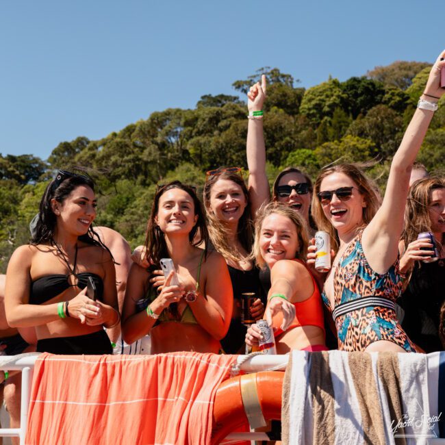 A group of women in swimwear are gathered together outdoors, smiling and holding drinks with trees and a blue sky in the background. Some towels and clothing are draped over a railing in front of them, capturing the perfect moment at a Sydney boat party hire.