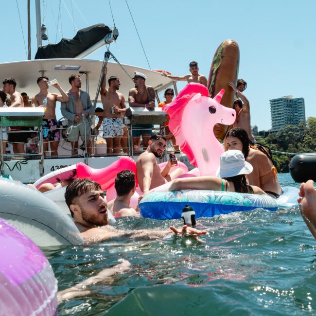 A group of people is enjoying a lively day on a boat and in the water. They are surrounded by inflatable pool floats, including a large unicorn. Some are in swimwear on the boat, while others are playing in the water with aqua jets. Trees and a tall building are visible in the background.