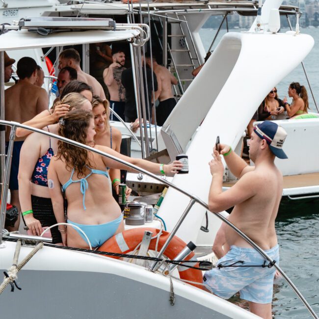 A group of people gather on a boat, some wearing swimwear and others casually dressed. The boat is surrounded by water, with another boat visible in the background. One person captures a photo of two women posing, while others mingle and chat near the calm waves.
