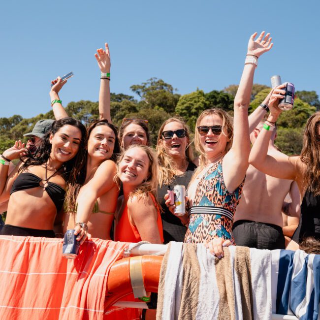 A group of people in swimwear pose and smile on a catamaran party Sydney boat, holding drinks and making celebratory gestures with a backdrop of trees and a clear blue sky.