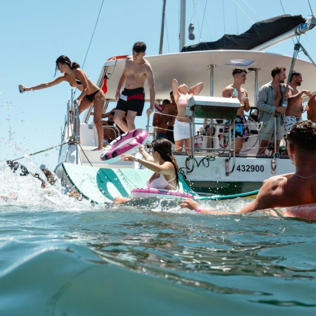 A group of people enjoy a sunny day on a boat and in the water. Some are on the deck, while others float using inflatable rings. One person is pouring water from a bottle into the sea. The boat buzzes with excitement and laughter, with clear skies and shimmering waters in the background.