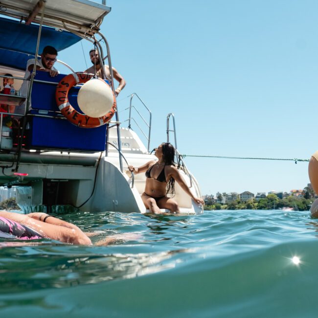 People enjoying a sunny day on a boat and in the water. A woman swims near the boat’s edge, another climbs a ladder back onto the boat, while others relax on inflatables and the boat deck. The background shows clear blue skies and distant buildings. Logos for The Yacht Social Club and Sail Gustavo are visible.