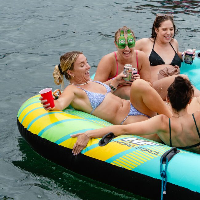 Four women are lounging on a large inflatable raft on the water, smiling and enjoying drinks. One woman has cucumber slices on her eyes, adding a playful touch. The brightly colored raft with yellow and green stripes contrasts beautifully against the serene water backdrop, creating a picture-perfect scene.
