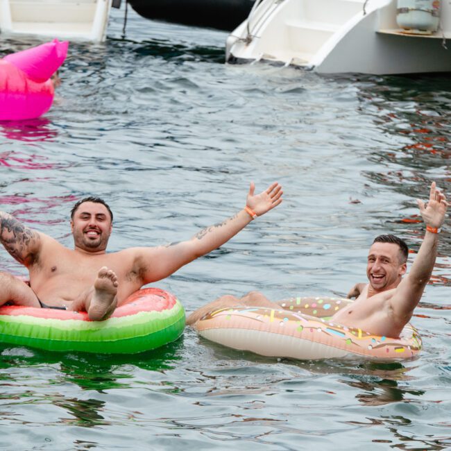 Two men float in the water on inflatable rings, one shaped like a watermelon slice and the other like a donut. Both have their arms raised, smiling and enjoying their time. A boat and a bright pink flamingo floatie are visible in the background.