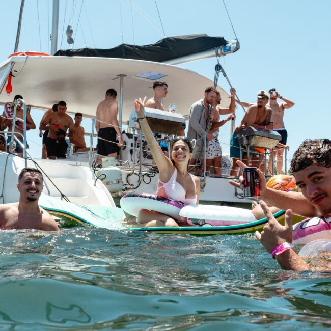 A lively group of people enjoying a sunny day on a boat and in the water. Several are lounging on inflatables, holding drinks, and engaging in casual conversation. The boat and a bright, clear sky are in the background. The atmosphere is festive and joyful.
