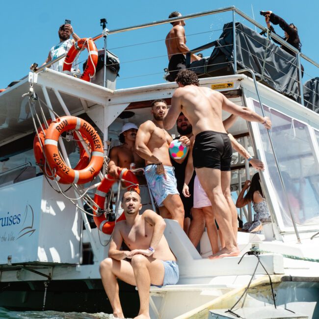A group of friends enjoying a sunny day on a boat. Some are standing on the deck, chatting and holding a vibrant beach ball, while others sit on the edge with feet in the calm, clear water. Lifebuoys adorn the railings. The atmosphere is filled with laughter.