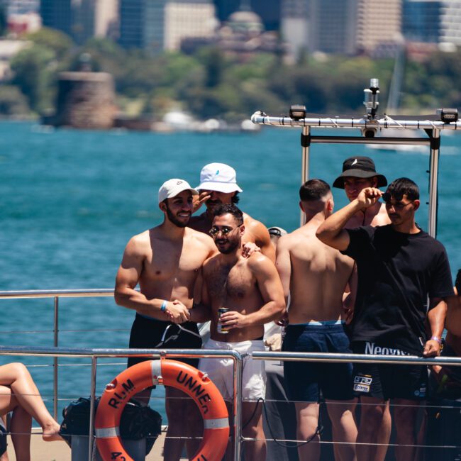 A group of people, mostly shirtless men, stand and chat on the deck of a boat with lifebuoys labeled “PM UNICAP.” The boat is on a body of water with a cityscape in the background. Two logos reading “Sail Gustavo” and “Leandro Soares” are in the bottom corners, highlighting a lively atmosphere.