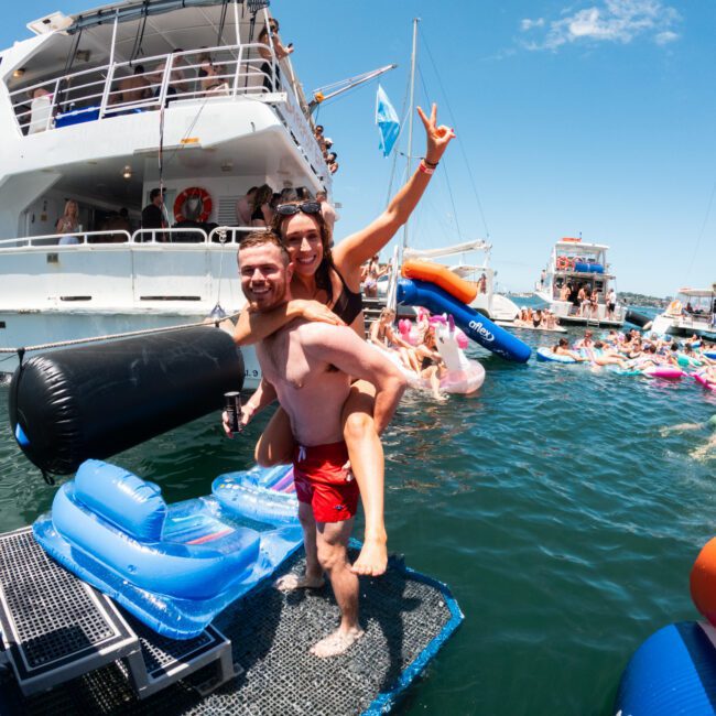 A man in red swim trunks carries a woman in a bikini on his back while standing on a floating platform near a boat. They're both smiling and making peace signs. The surrounding water is filled with colorful inflatables and other people enjoying the vibrant summer day.
