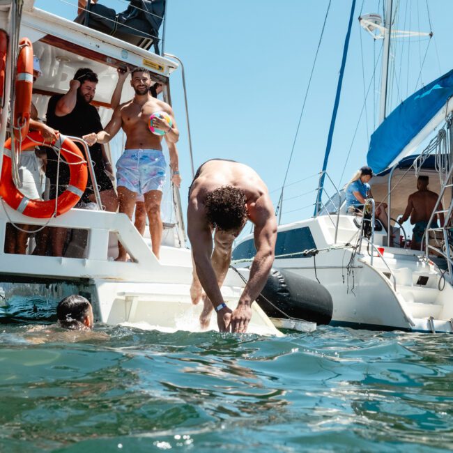 A man dives into the water from a boat while others stand and watch. Another person surfaces nearby. Two sailboats with blue canopies are visible, and people are enjoying the sunny day on deck. Life rings are attached to the boats, and the calm waters shimmer under the radiant sky.