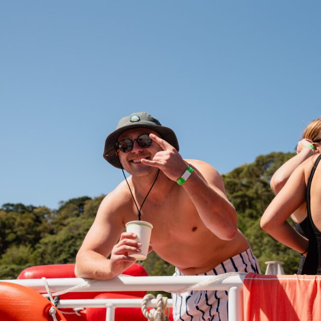 A shirtless man wearing a hat and sunglasses holds a cup while making a gesturing hand sign on a boat with several people. Trees and blue sky are in the background, capturing the fun of a Sydney boat party hire.