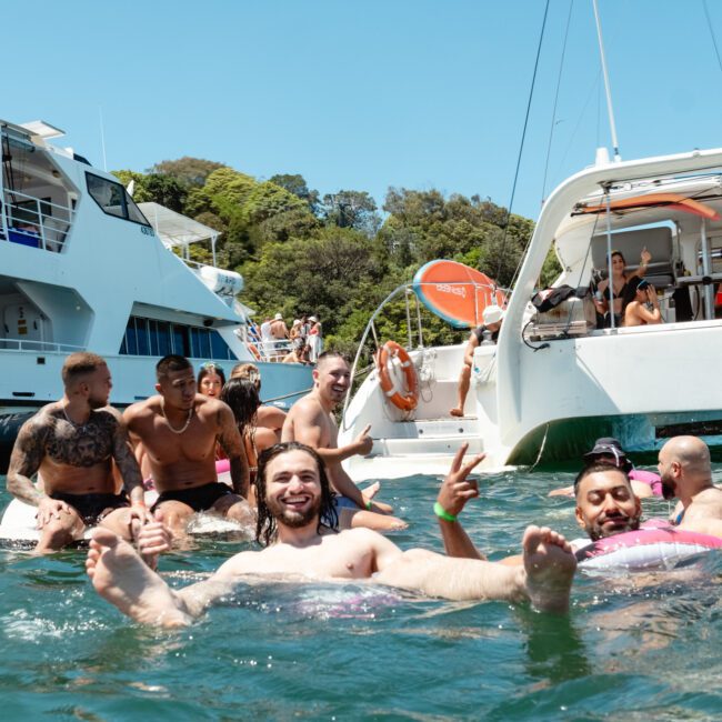A group of people enjoying a sunny day in the water near two boats. Some are relaxing on inflatable floaties while others are swimming and socializing with vibrant music playing in the background. The scene is lively with people smiling and showing peace signs. Trees and a clear blue sky frame the idyllic setting.