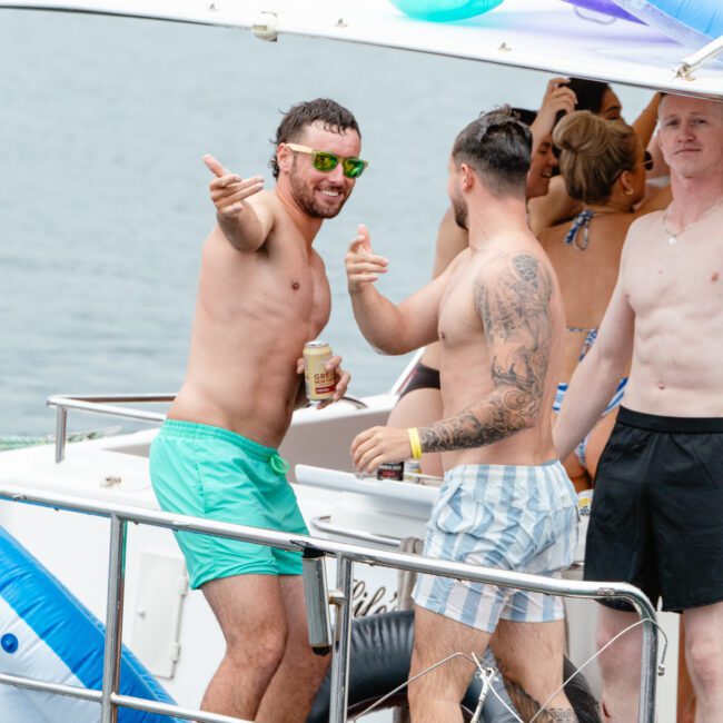 A group of people enjoying a boat party. Two shirtless men are at the foreground, one wearing sunglasses, green shorts, and pointing while holding a drink. The other man, heavily tattooed and wearing striped shorts, is facing him. Others are visible in the background as they socialize under the sun.