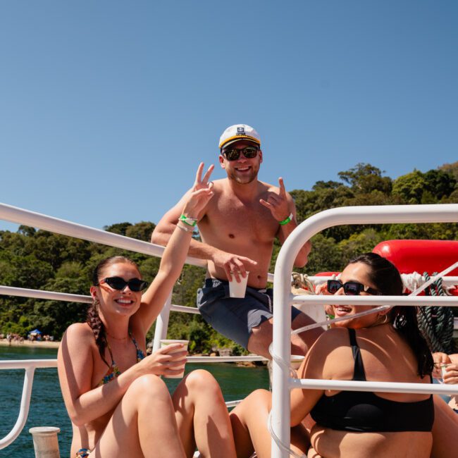 Three people in swimwear enjoying time on a luxury yacht with one person giving a peace sign and holding a drink. Trees and clear blue sky are in the background.